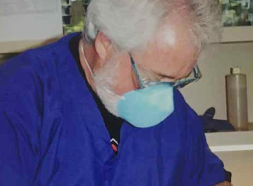 The image shows a man wearing a blue surgical gown and a face mask, sitting at a desk with his head down, appearing to be working on something.