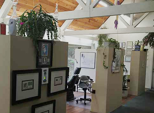The image depicts an office space with a beige carpeted floor, white walls, and various pieces of furniture and decorations.