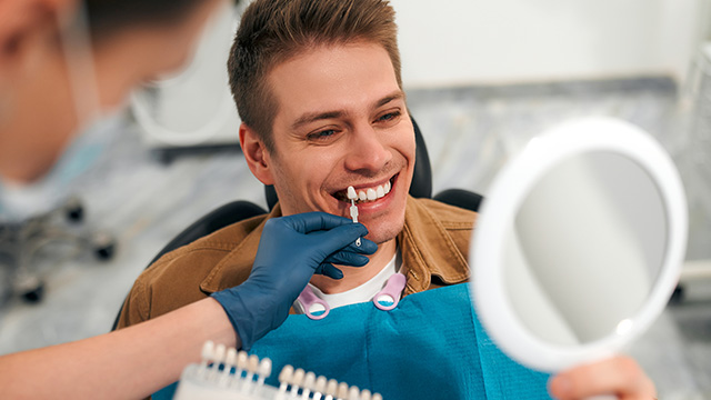 The image shows a man sitting in a dental chair with his mouth open, receiving dental care from a professional who is using a mirror to examine his teeth.