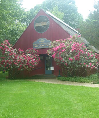 The image shows a red building with a sign that reads The Old School House surrounded by blooming pink flowers, set against a backdrop of a clear sky.